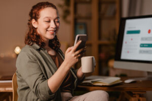 Mulher sorridente usando smartphone em casa, com computador e caneca na mesa, representando produtividade e bem-estar no ambiente de trabalho digital.